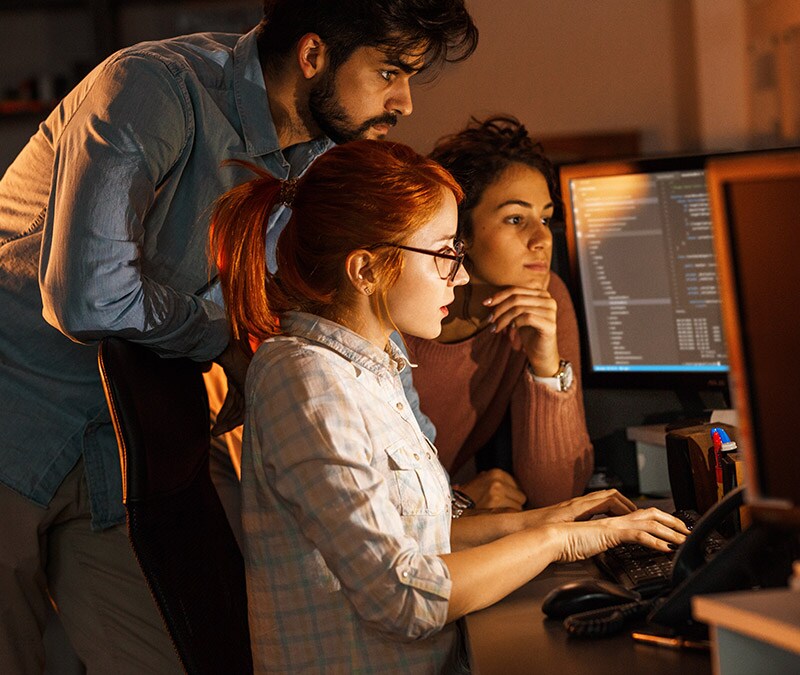 Three young adults looking at a computer monitor together, reading an article about the most famous computer viruses.