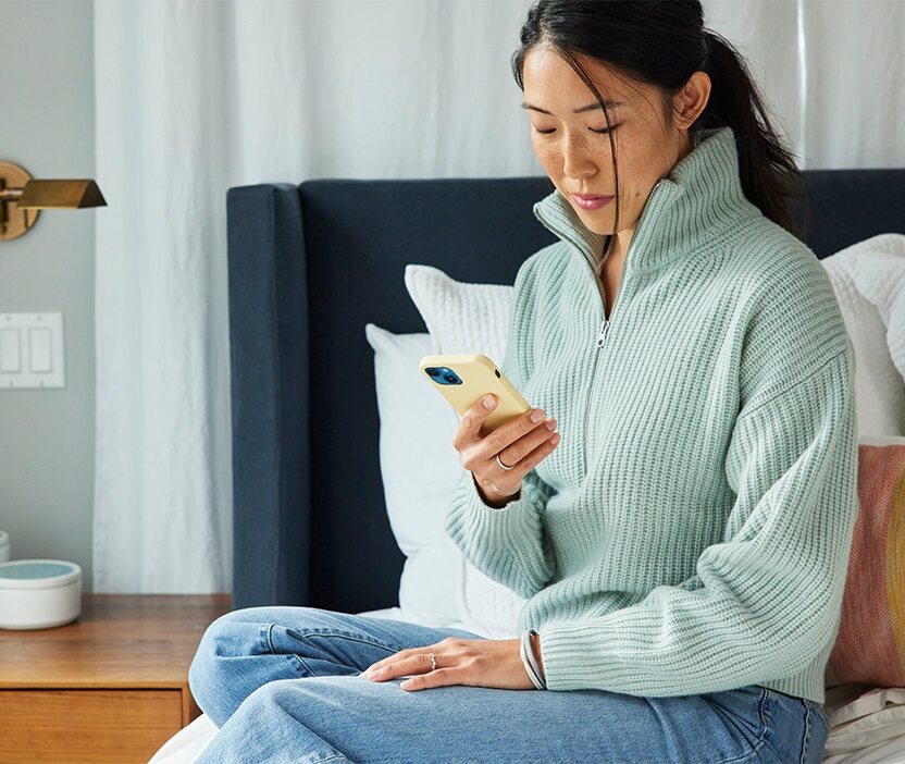 A woman sits on a bed, looking down at her phone and thinking about how to create a memorable password.