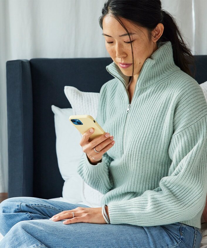 A woman sits on a bed, looking down at her phone and thinking about how to create a memorable password.